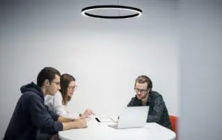 A corporate photograph featuring three colleagues working together at a round table with a laptop and notepads, under a modern circular light fixture in a Sydney office.