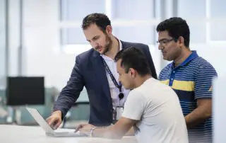 A group of three colleagues collaborating in an office, with a man in a suit pointing at a laptop while two others observe. Corporate photography by Sydney photographer Gavin Jowitt.