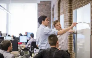 A group of colleagues collaborating in an open-plan office with exposed brick walls. Two men stand at a whiteboard, pointing and discussing ideas, while others work at desks with computer monitors in the background. Captured by Sydney corporate photographer Gavin Jowitt.