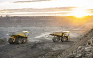 industrial photograph showcasing two large yellow mining haul trucks parked on a rugged mining site at sunrise, with soft light highlighting the quarry landscape.