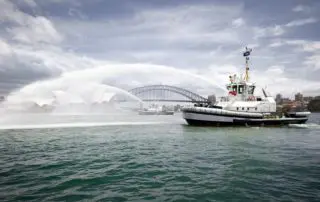 A marine photograph capturing a tugboat spraying water jets in Sydney Harbour with the Sydney Opera House and Harbour Bridge visible in the background.