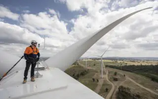 Photograph of a technician in safety gear standing on top of a wind turbine, with expansive views of a wind farm and rolling hills under a partly cloudy sky in Australia.